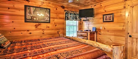 Un dormitorio tipo cabaña de troncos, con cama, ventilador de techo, televisor y paredes de madera.
