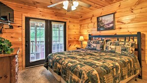 Une chambre dans une cabane en bois, avec un lit, un ventilateur de plafond, une porte avec des panneaux de verre et un tableau encadré au mur.