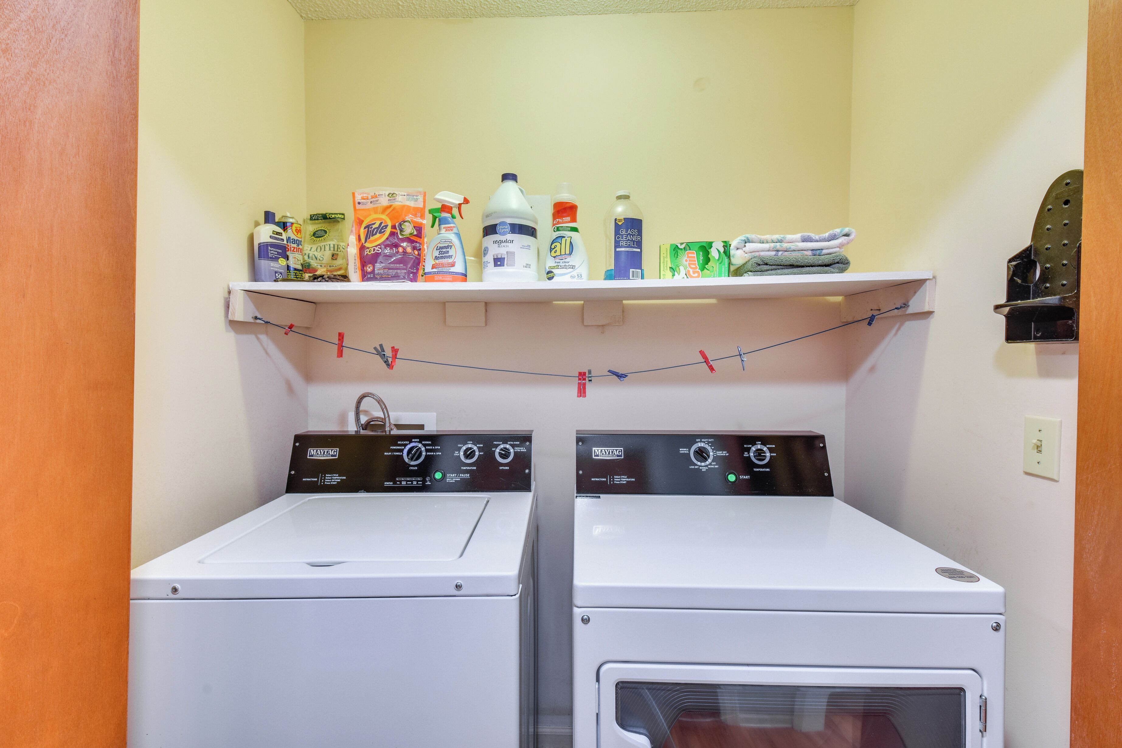 Une buanderie équipée de deux machines à laver à chargement frontal et d’un sèche-linge, une étagère au-dessus pour ranger divers produits de nettoyage, et une porte avec un porte-serviettes.