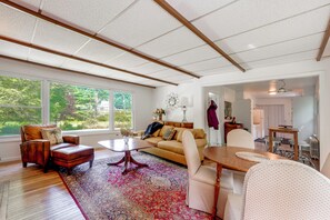 A spacious living room with a large window, a sofa, a coffee table, and a dining area with chairs. - Spacious Sullivan County House w/ Fire Pit (Hurleyville)