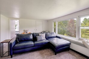 A room with a dark leather sofa, a small wooden table, and a view of trees through a large window. - Spacious Sullivan County House w/ Fire Pit (Hurleyville)