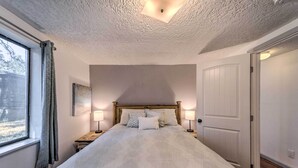 A bedroom with a large bed, a wooden headboard, two bedside lamps, a window with curtains, a door, and a ceiling light fixture. - Ruidoso Cabin - Walk to Midtown Dining & Shops! (Ruidoso)