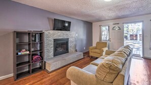 A living room with a fireplace, a sofa, a bookshelf, and a television. - Ruidoso Cabin - Walk to Midtown Dining & Shops! (Ruidoso)