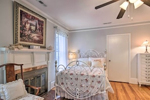 Un dormitorio con estructura de cama de metal blanco, chimenea, un cuadro y cómoda blanca.