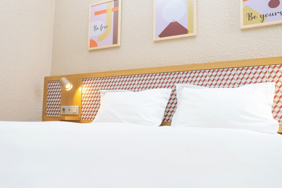 A hotel room with a bed, pillows, a nightstand, a lamp, and wall art.