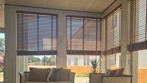 A modern interior with a beige sofa, a round coffee table, and a white armchair. Large windows with horizontal blinds allow natural light. - Flag Hotel Lisboa Oeiras (Oeiras)