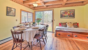 A dining area with a wooden table set for four, a window with a view of trees, and a bed with a floral pillow. - Old Time Catskill Mtn Retreat, Handcrafted Cabin! (East Durham)