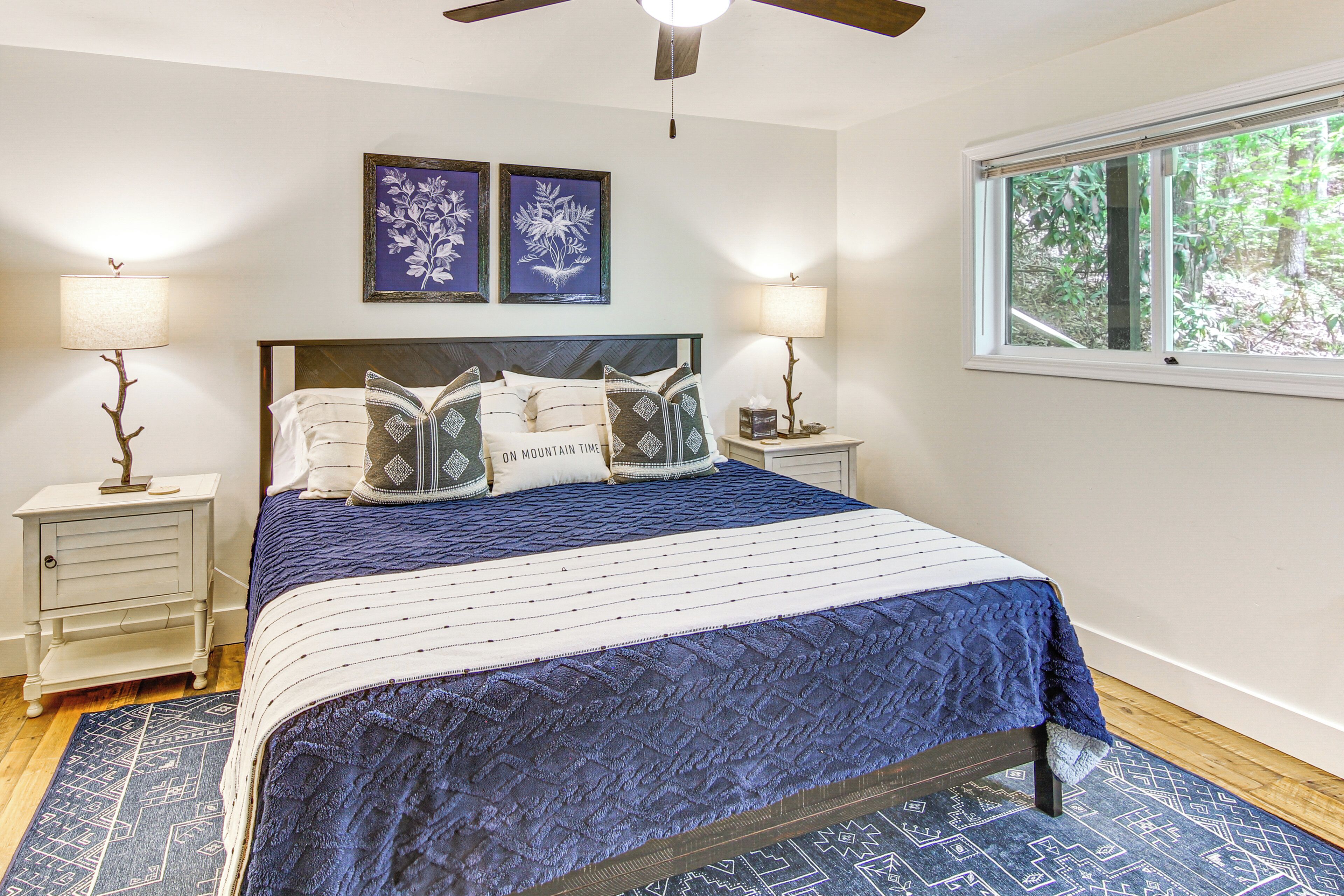 A bedroom with a bed, two bedside tables, a nightstand, a ceiling fan, and a window with a view of trees.