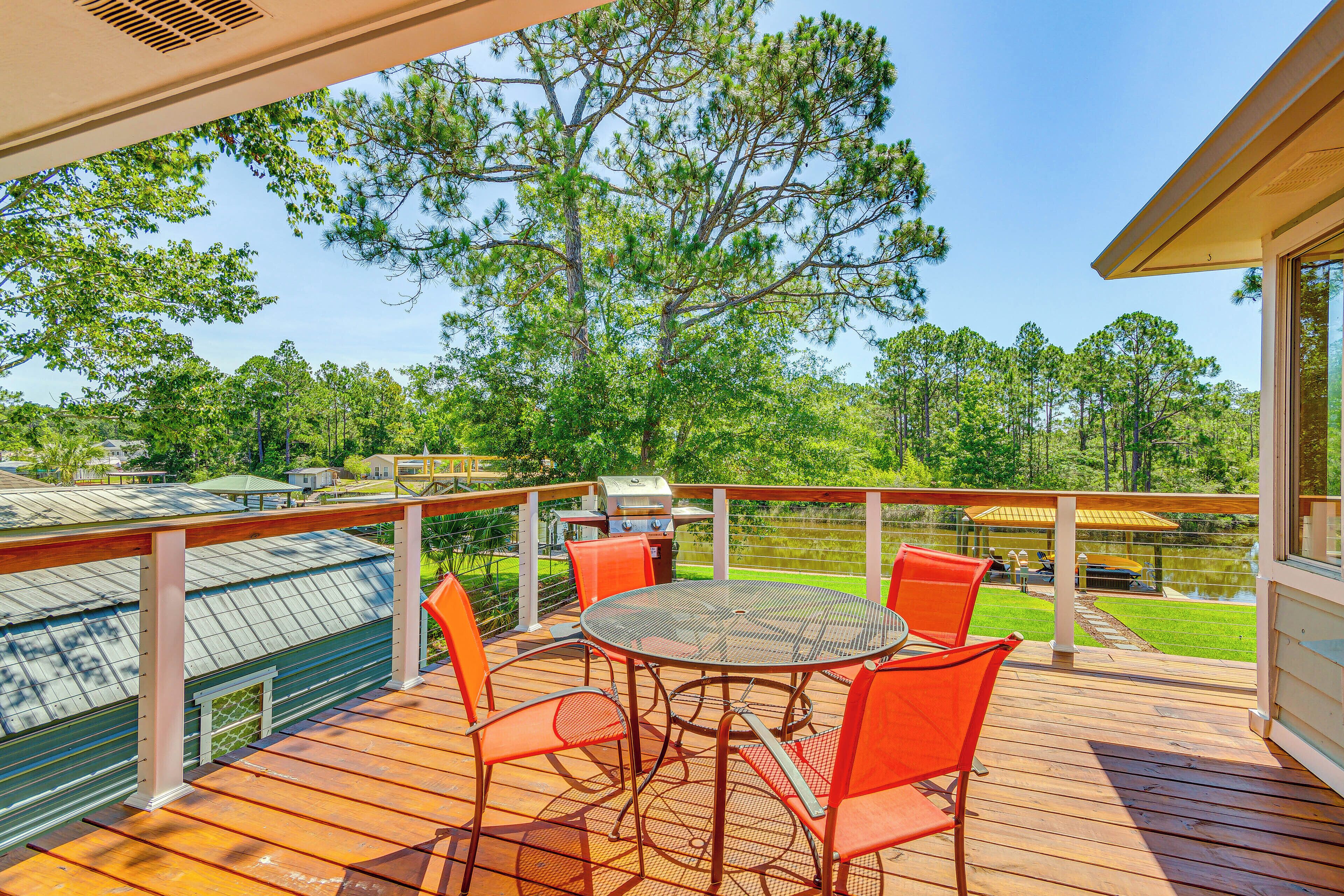 A wooden deck with a round table and orange chairs, overlooking a lake and trees.