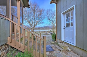 Un escalier en bois mène à une maison dont les fondations sont en pierre, avec une porte blanche et une vue sur un lac et des arbres.