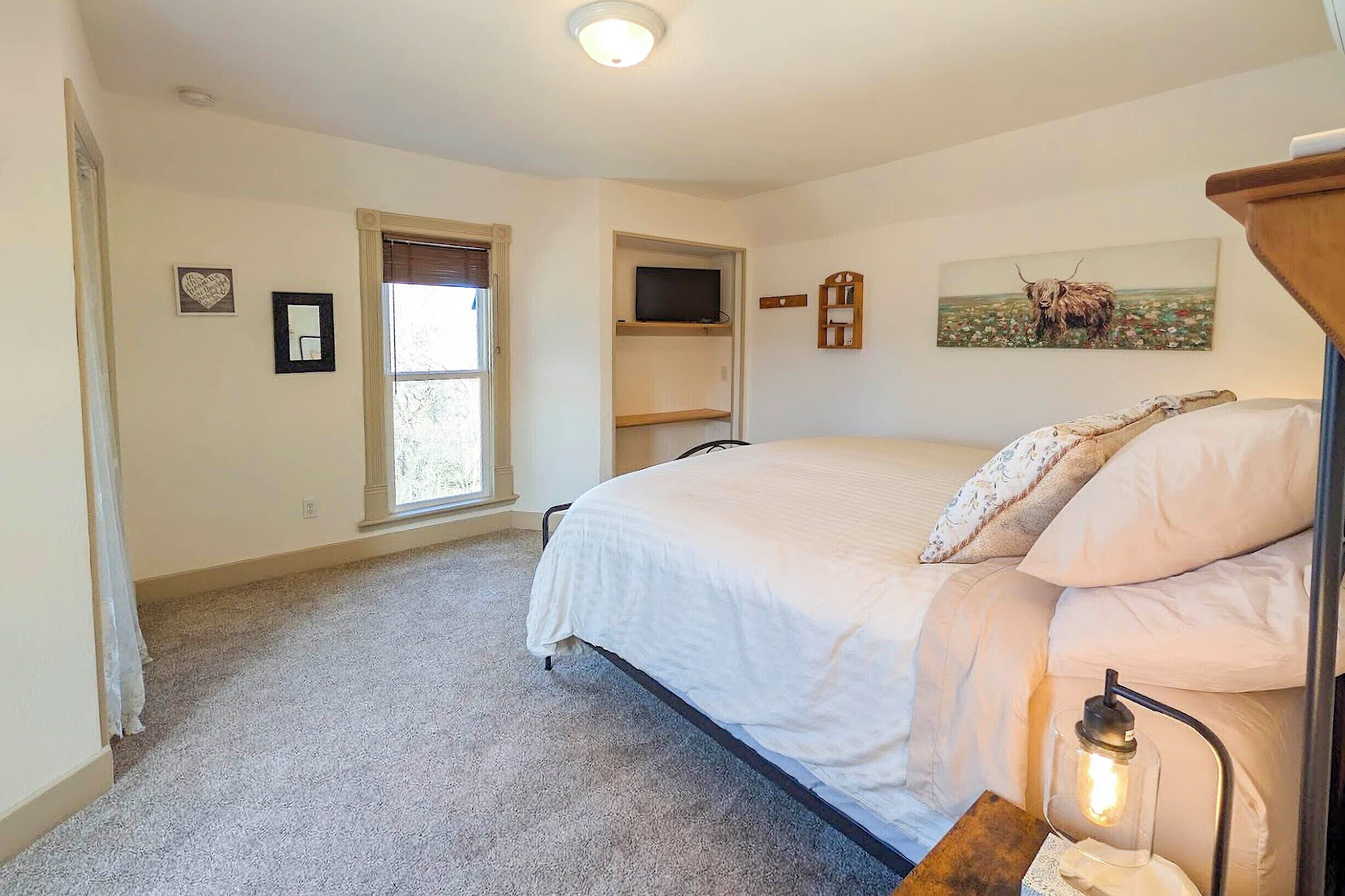 A bedroom with a bed, a window, a TV shelf, and a painting on the wall.