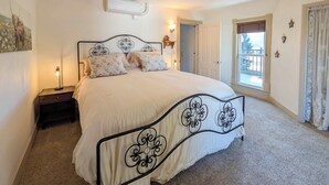 A bedroom with a large bed, a nightstand, a wall-mounted air conditioner, a window with curtains, and a door. - Charming Stevensville Home w/ Open Yard! (Stevensville)