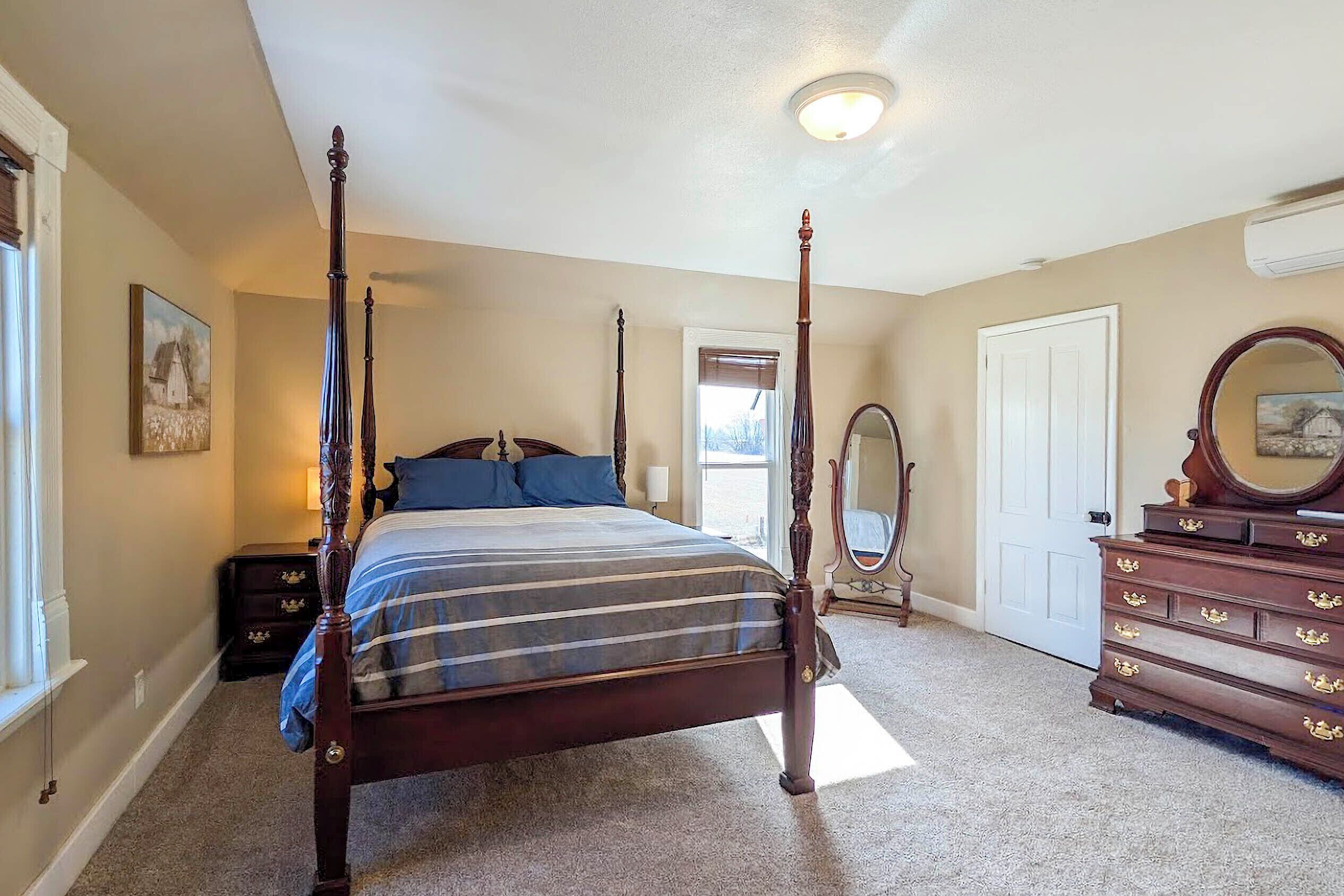 A four-poster bed with a striped bedspread, a dresser with drawers, a mirror, and a painting on the wall.