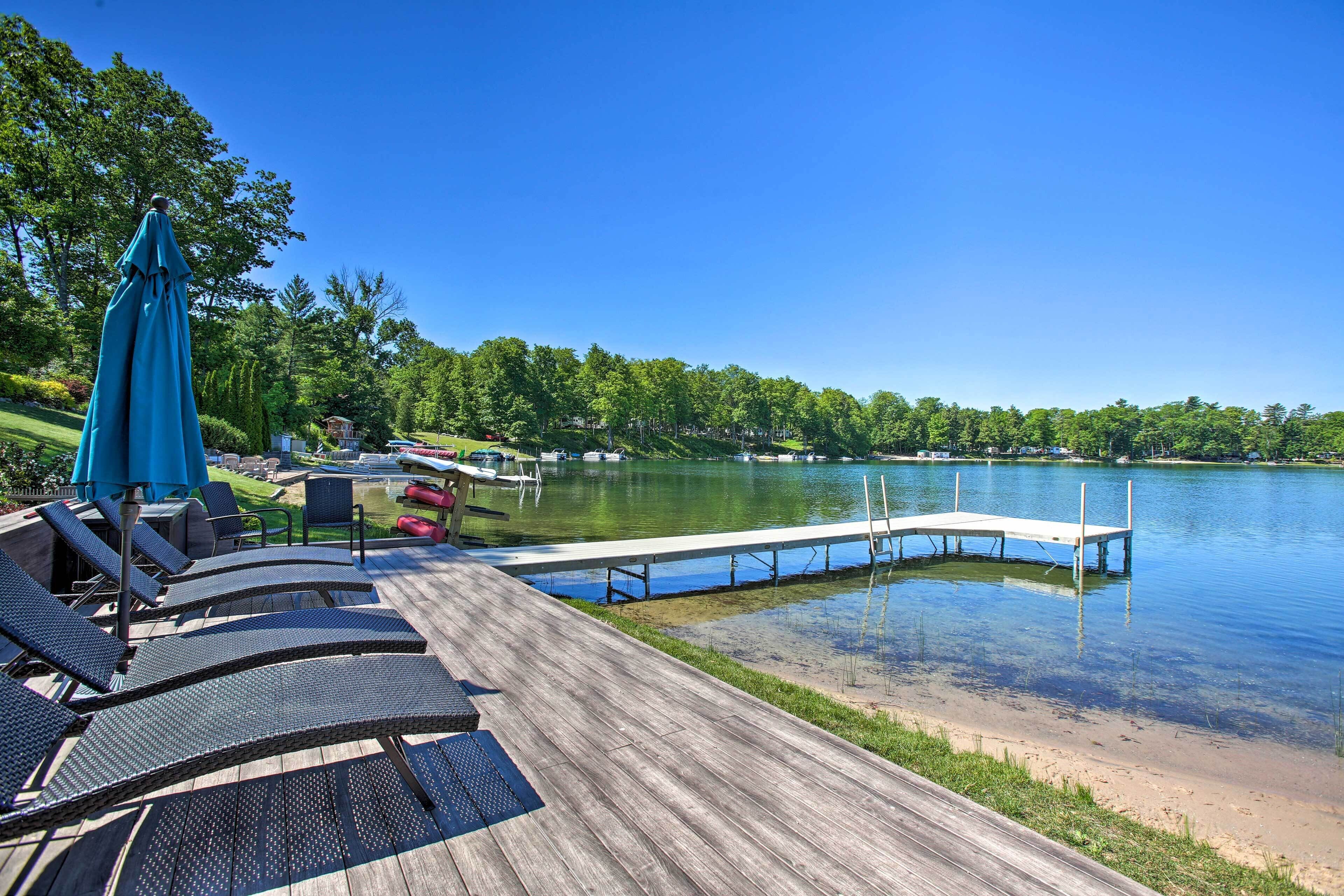 Une terrasse en bois avec des chaises longues et un parasol bleu, donnant sur un lac paisible avec une jetée qui s’avance dans l’eau.