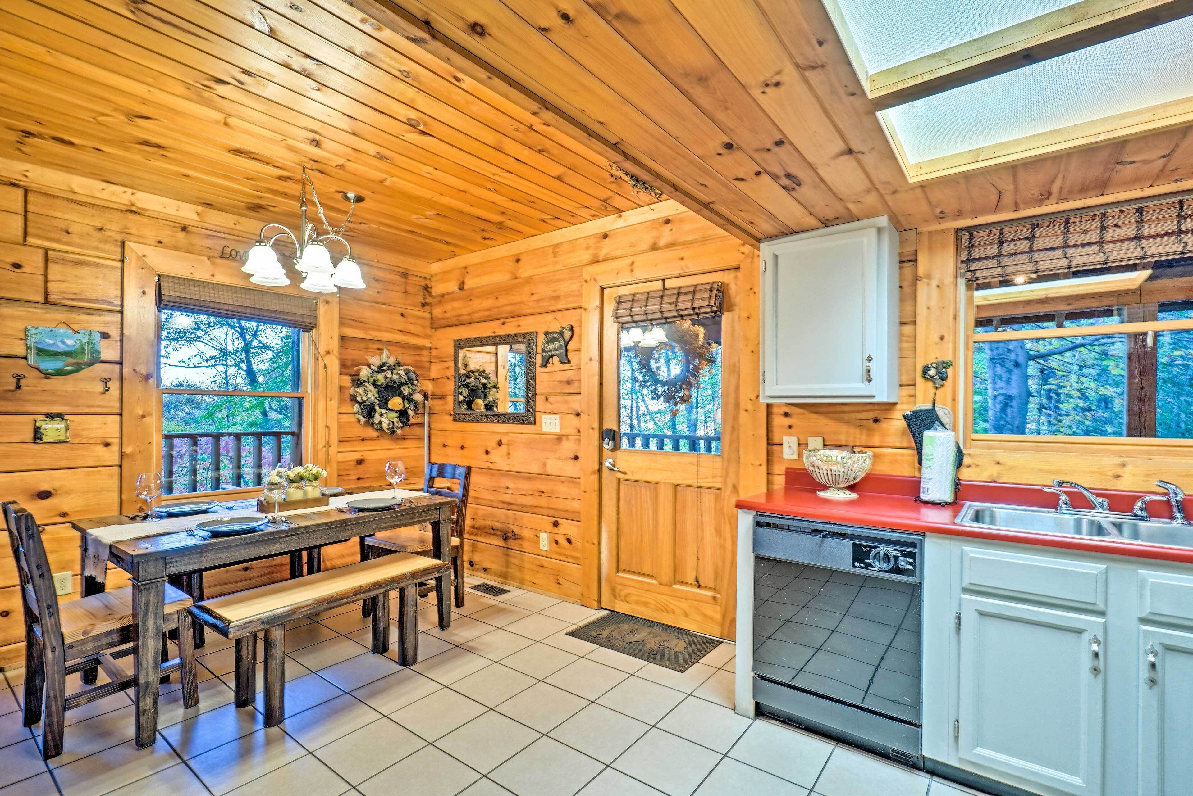 A wooden cabin interior with a dining table, benches, a kitchen with a red countertop, and a large window.