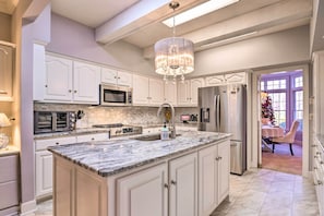 A spacious kitchen with white cabinetry, a large island, a chandelier, and a view to a dining area with a Christmas tree.