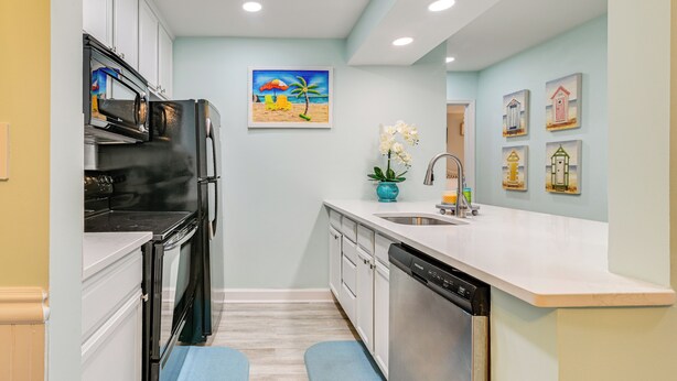 A modern kitchen with white cabinets, a black refrigerator, a dishwasher, and a sink. There is a painting of a beach scene on the wall and a vase with flowers on the counter. - Cozy Myrtle Beach Getaway: 1 Block to Ocean! (Myrtle Beach)