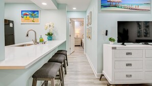 A kitchen with a white countertop, a sink, and bar stools. There is a TV on a white cabinet and a painting of a beach scene on the wall. - Cozy Myrtle Beach Getaway: 1 Block to Ocean! (Myrtle Beach)