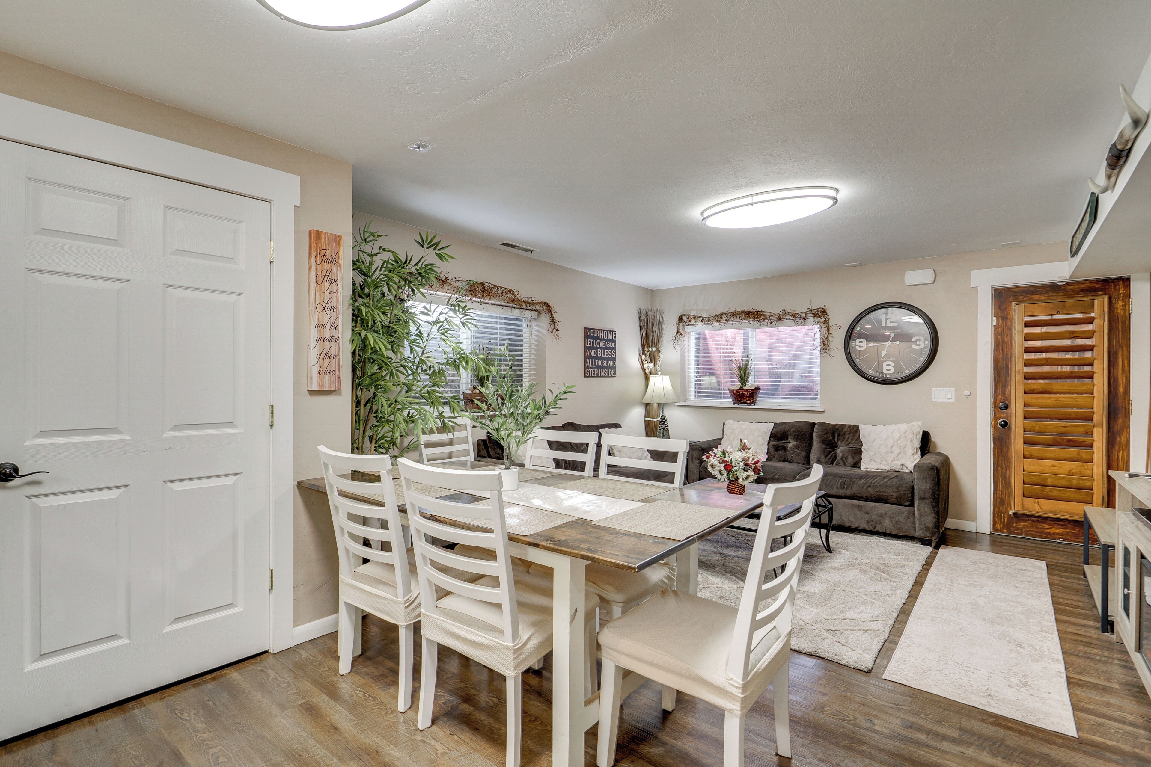 A dining area with a wooden table and white chairs, a sofa, and a clock on the wall.