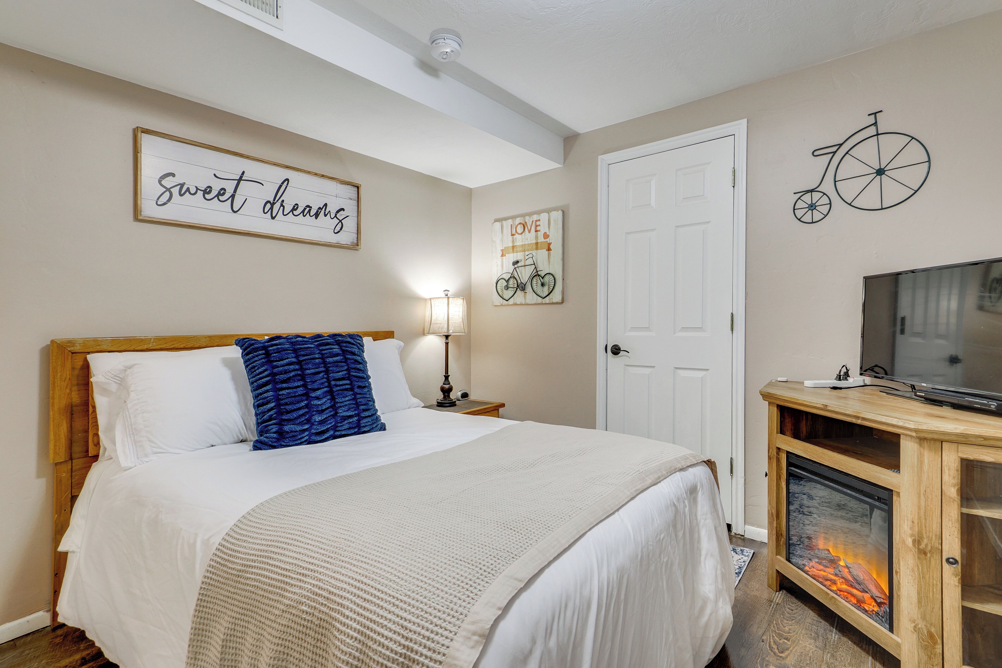 A neatly arranged bedroom with a bed, a television on a wooden stand, a fireplace, and wall decorations including a bicycle and a sign that says "sweet dreams".