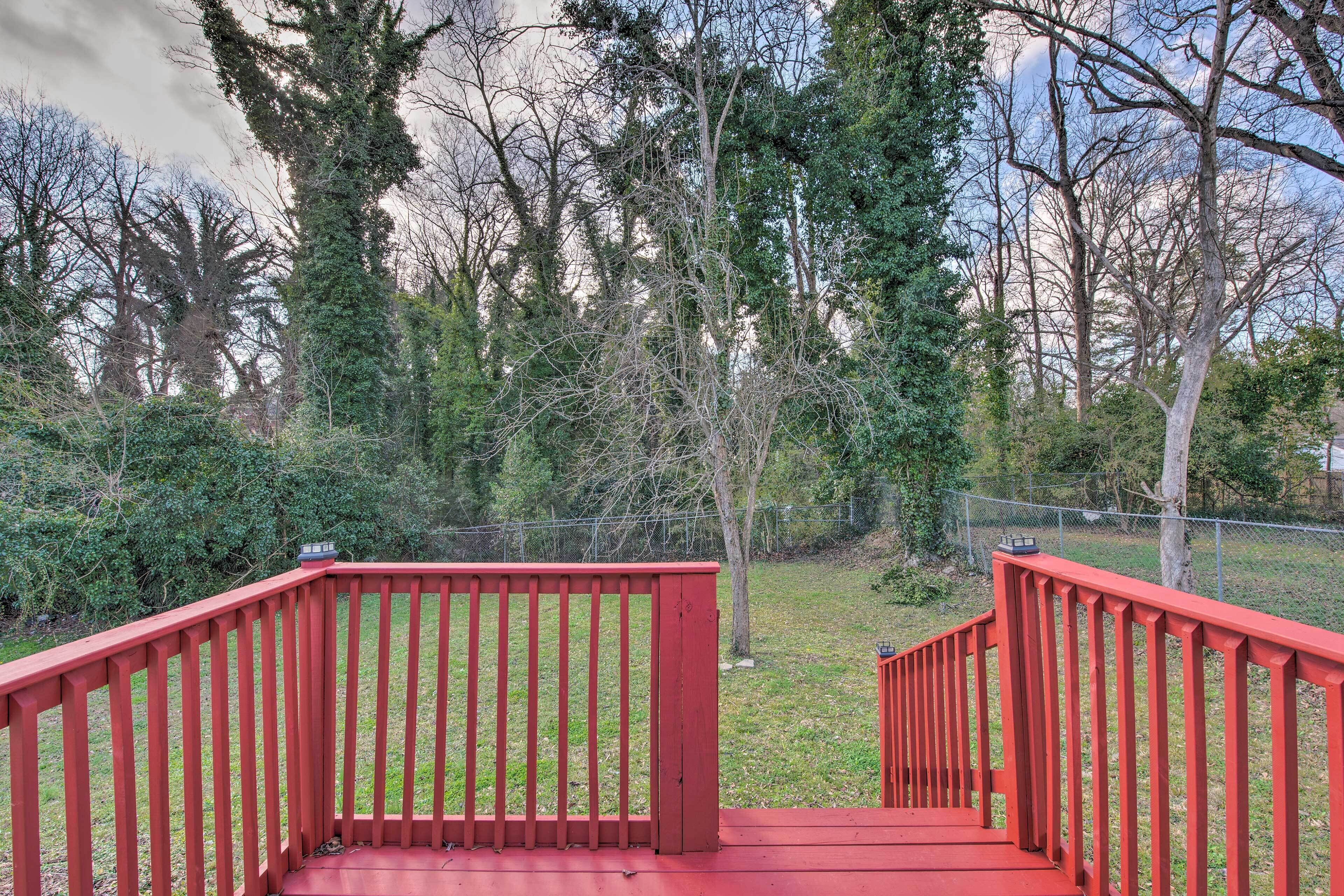 A red wooden staircase leading to a deck with a metal railing, surrounded by trees and a grassy yard.