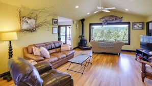 A living room with a leather sofa, a coffee table, a fireplace, and a large window with a view of trees. - Hermosa Home w/ Black Hills View, Gas Grill & Deck (Hermosa)