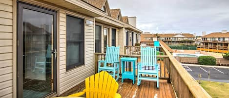 A wooden deck with yellow and blue Adirondack chairs, a small table, and a glass door leading to a covered area.