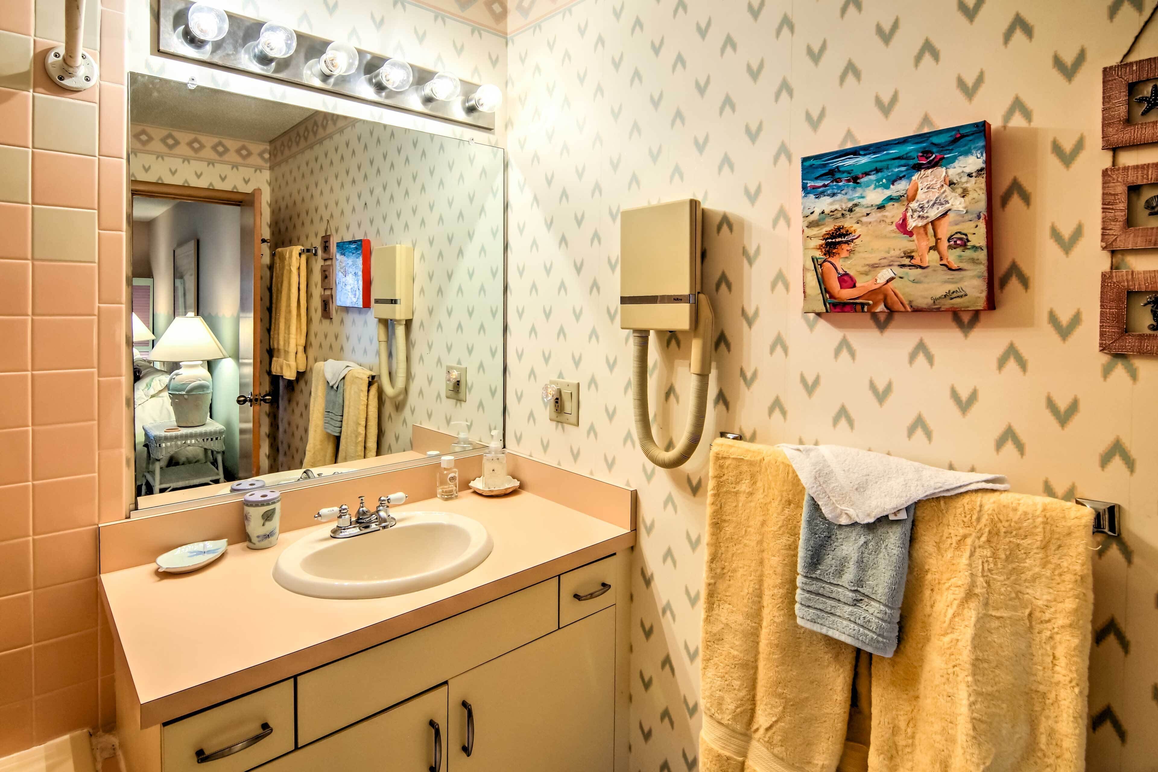 Une salle de bain équipée d’un lavabo, d’un miroir, d’un porte-serviettes et d’un tableau représentant une scène de plage.