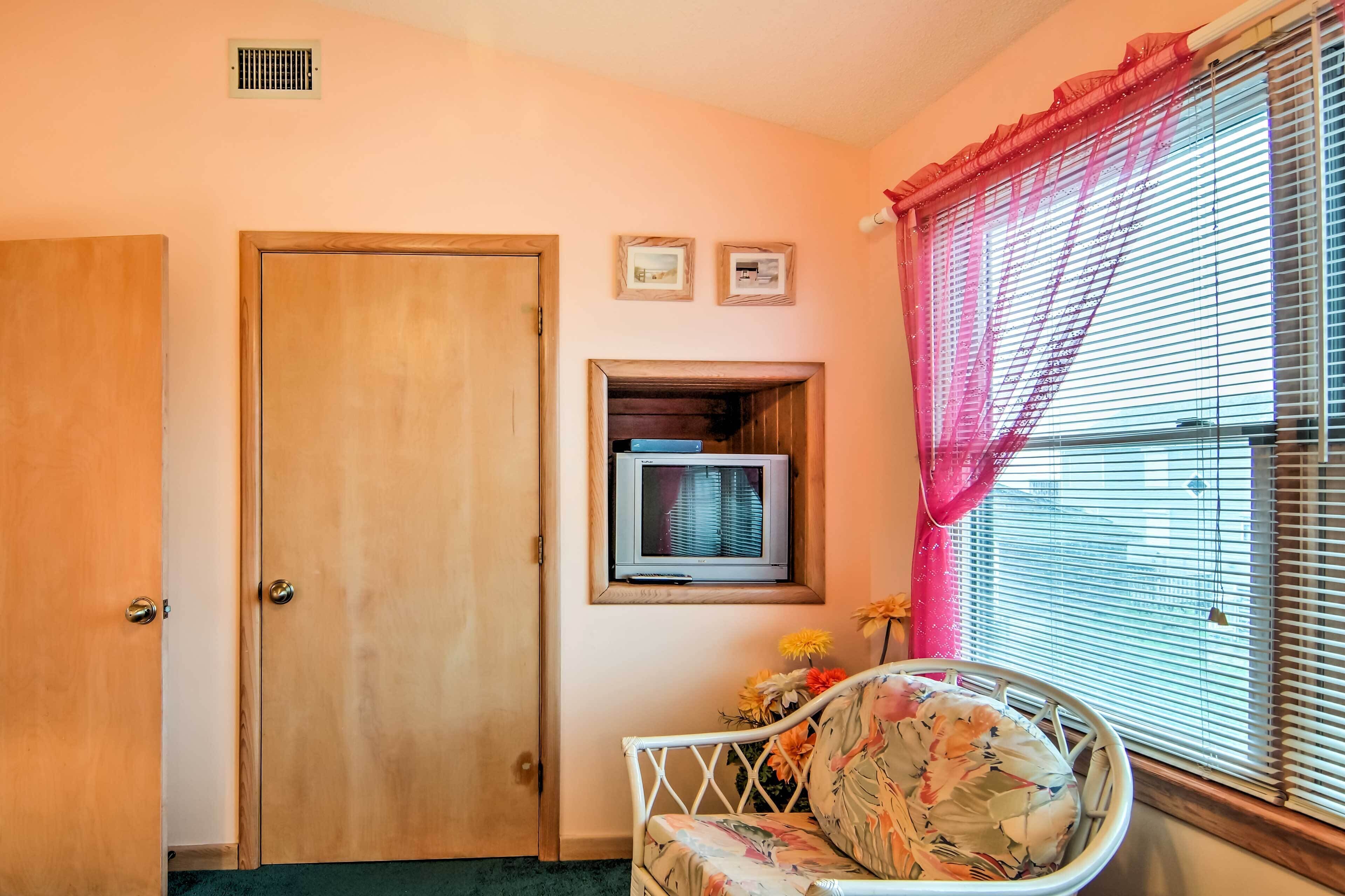 A room with a wooden door, a television mounted on the wall, a window with blinds, and a floral-patterned chair.