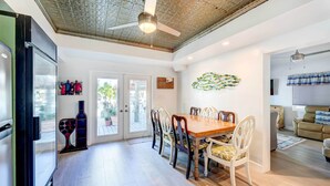 A spacious dining area with a wooden table and white chairs, a ceiling fan, and a decorative wall piece. - Canal-front Florida Keys Home w/ Dock! (Tavernier)