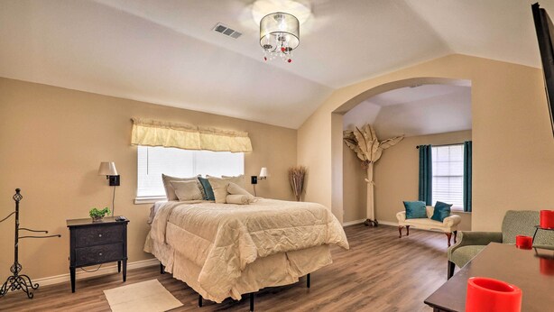 A bedroom with a bed, bedside tables, a lamp, a nightstand, a dresser, a window with curtains, a chair, a table, and a red candle.