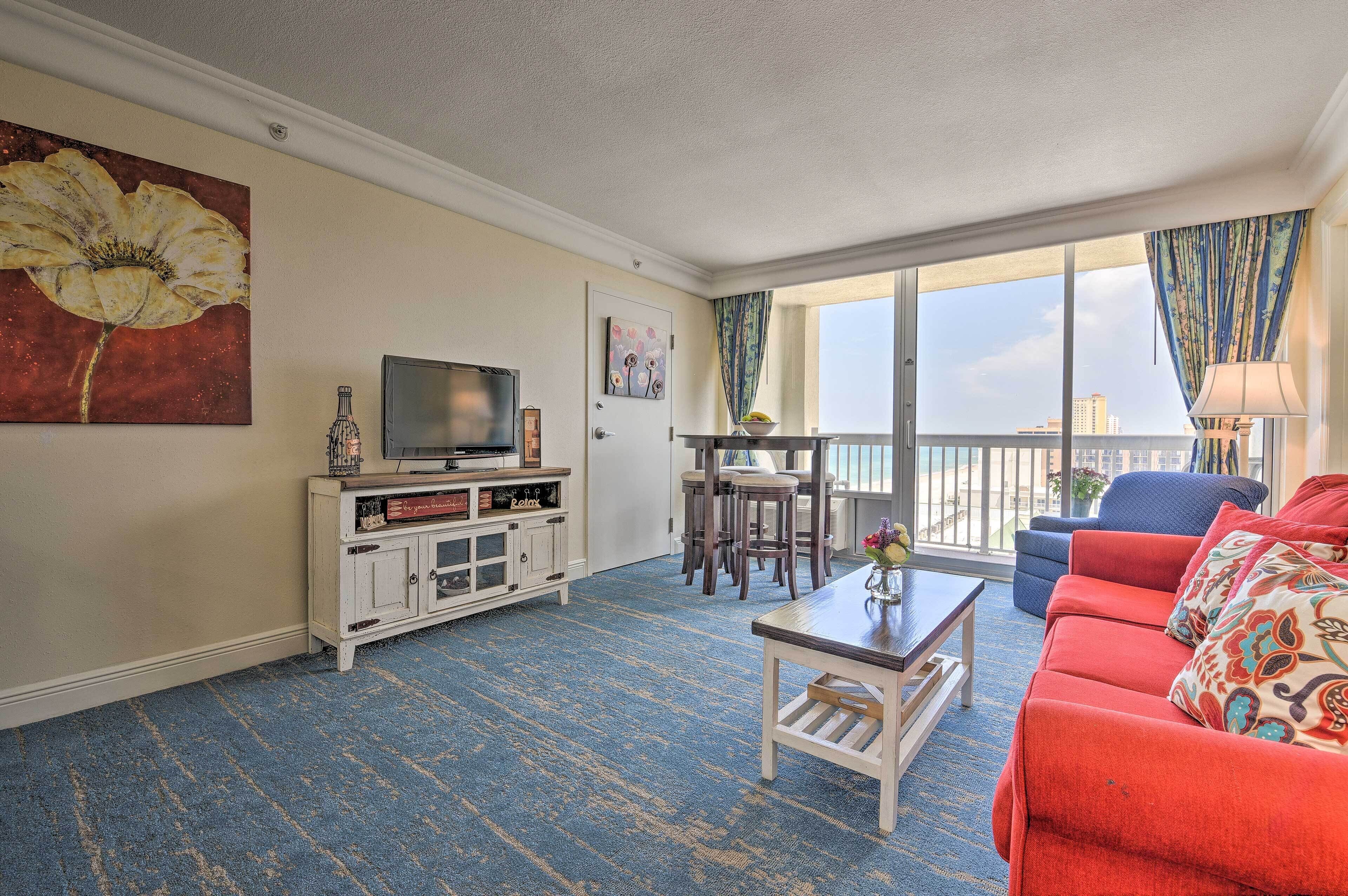 Una sala de estar con alfombra azul, un sofá rojo, un televisor y un balcón con vistas.