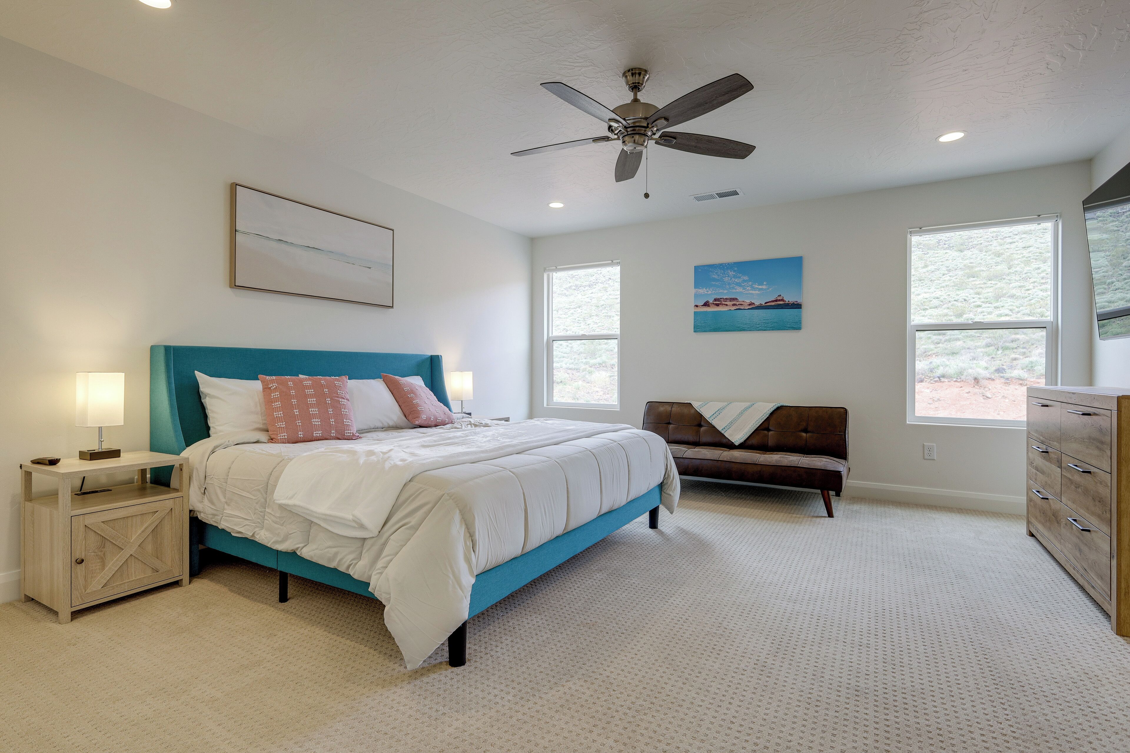 Une chambre à coucher comprenant un lit, une table de chevet, une commode, un ventilateur de plafond et deux fenêtres.