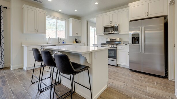 A modern kitchen with white cabinetry, a stainless steel refrigerator, a microwave, and a stove. There are three black bar stools at the island. - Sleek + Modern Home Rental Near Red Cliffs! (Washington)