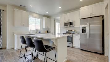 A modern kitchen with white cabinetry, a stainless steel refrigerator, a microwave, and a stove. There are three black bar stools at the island.
