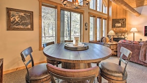 A dining area with a round wooden table, four chairs, a chandelier, and large windows with blinds. - Klamath Falls Cabin Retreat w/ Deck & Grill! (Klamath Falls)