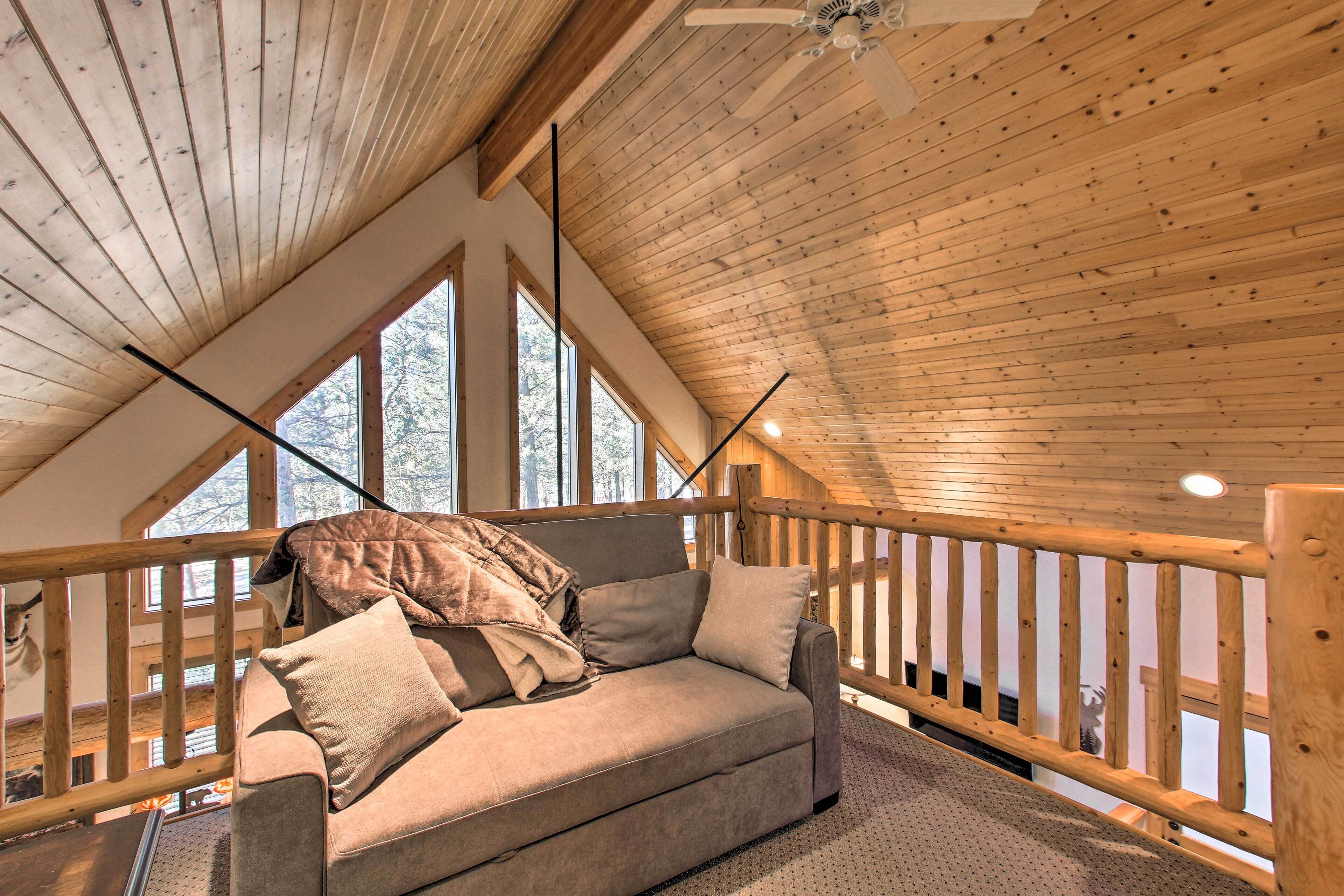 A cozy loft bedroom with a wooden ceiling, a cushioned sofa, and large windows offering a view of the outdoors.