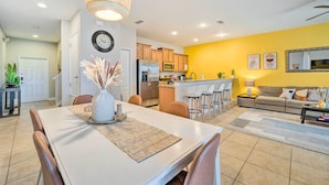 Uma cozinha moderna com eletrodomésticos de aço inoxidável, sala de jantar com mesa e cadeiras brancas e sala de estar com sofá e mesa de centro.