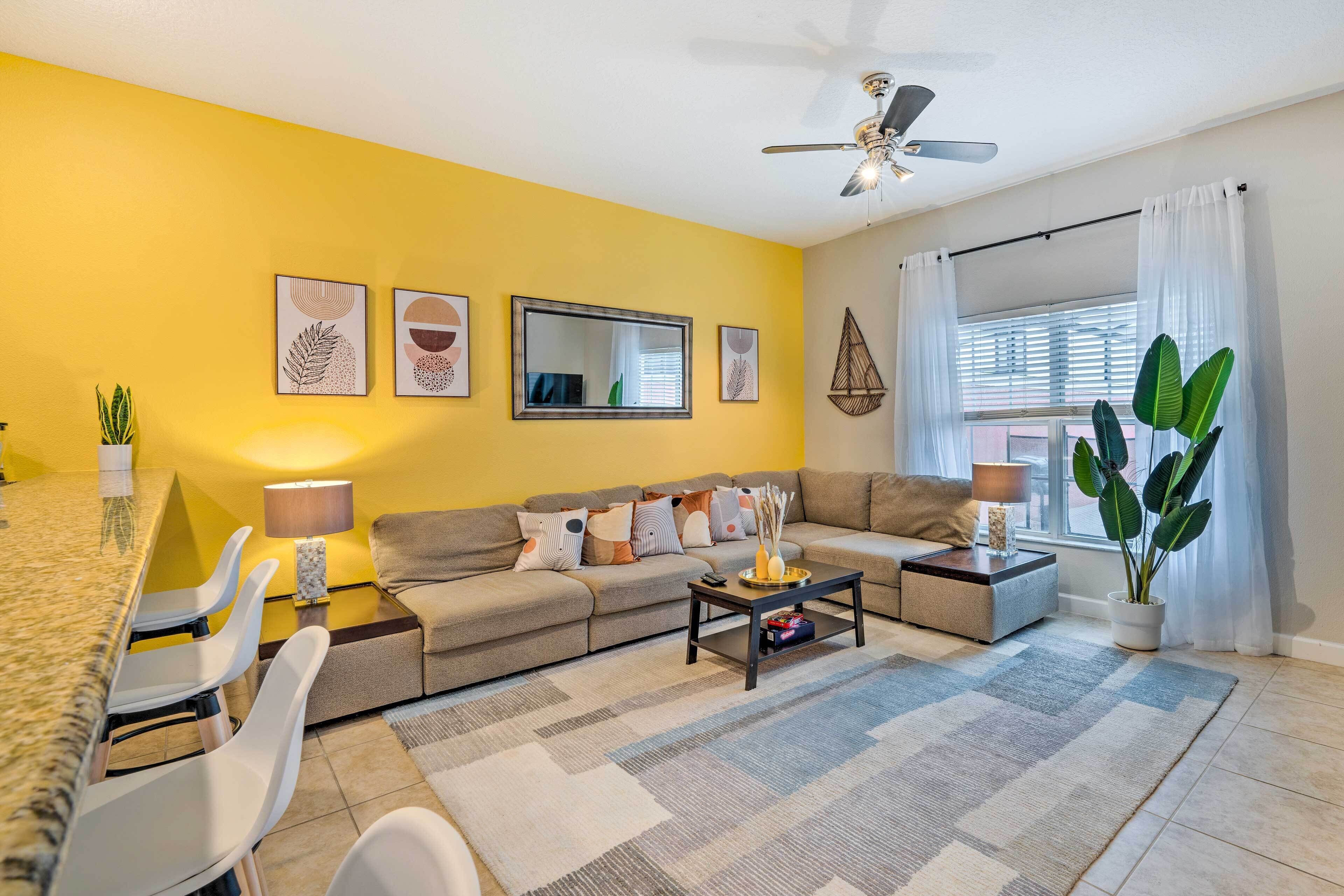A living room with a yellow wall, a ceiling fan, a sofa, a coffee table, and a dining area with chairs and a lamp.