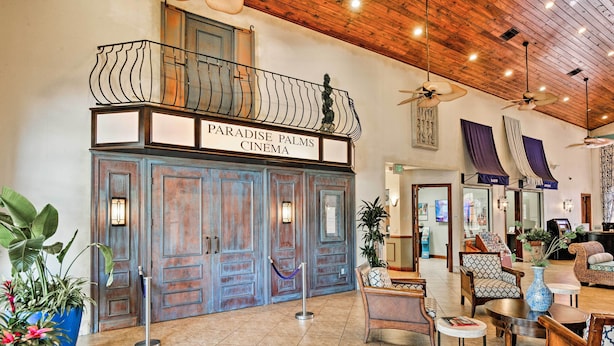 Um hall de entrada com teto de madeira, uma varanda com um letreiro indicando “Paradise Palms Cinema” e uma área de estar com diversas cadeiras e mesas.