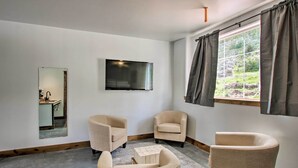 Un salon moderne doté d’un téléviseur à écran plat, de deux fauteuils beiges, d’une table basse en bois et d’une grande fenêtre agrémentée de rideaux.