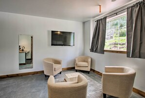Una sala de estar moderna con un televisor de pantalla plana, dos sillones color beige, una mesa de centro de madera y un ventanal con cortinas.