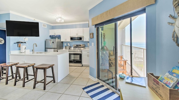 Una cocina moderna con armarios blancos, una isla grande con taburetes, un televisor de pantalla plana y vista al océano a través de una puerta corrediza de vidrio.