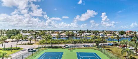 Un court de tennis comprenant deux terrains, entouré d’une clôture, avec vue sur un quartier résidentiel et un plan d’eau en arrière-plan.