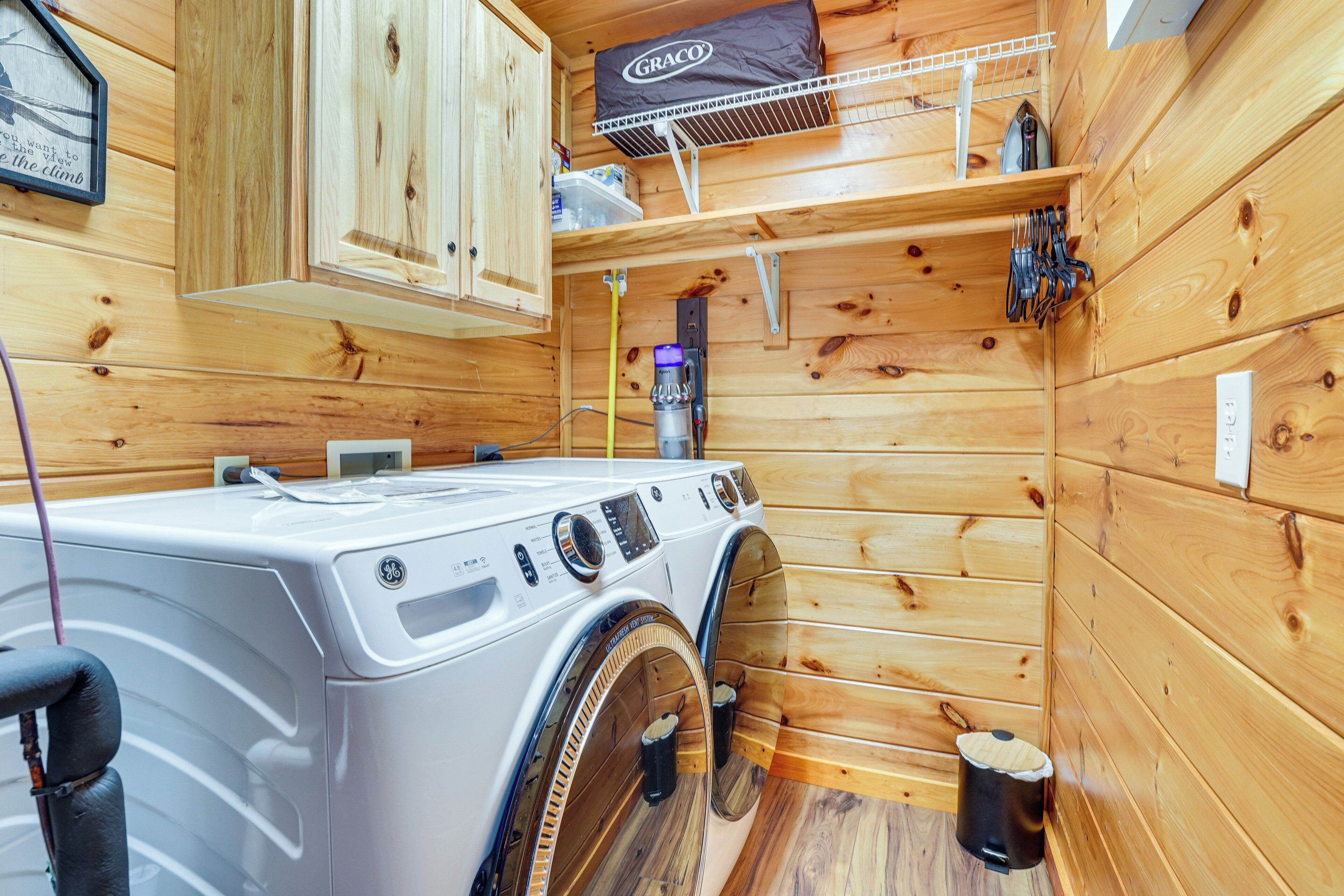 A compact laundry room with a front-loading washer and dryer, wooden walls, a small shelf with toiletries, and a hanging rack above the dryer.