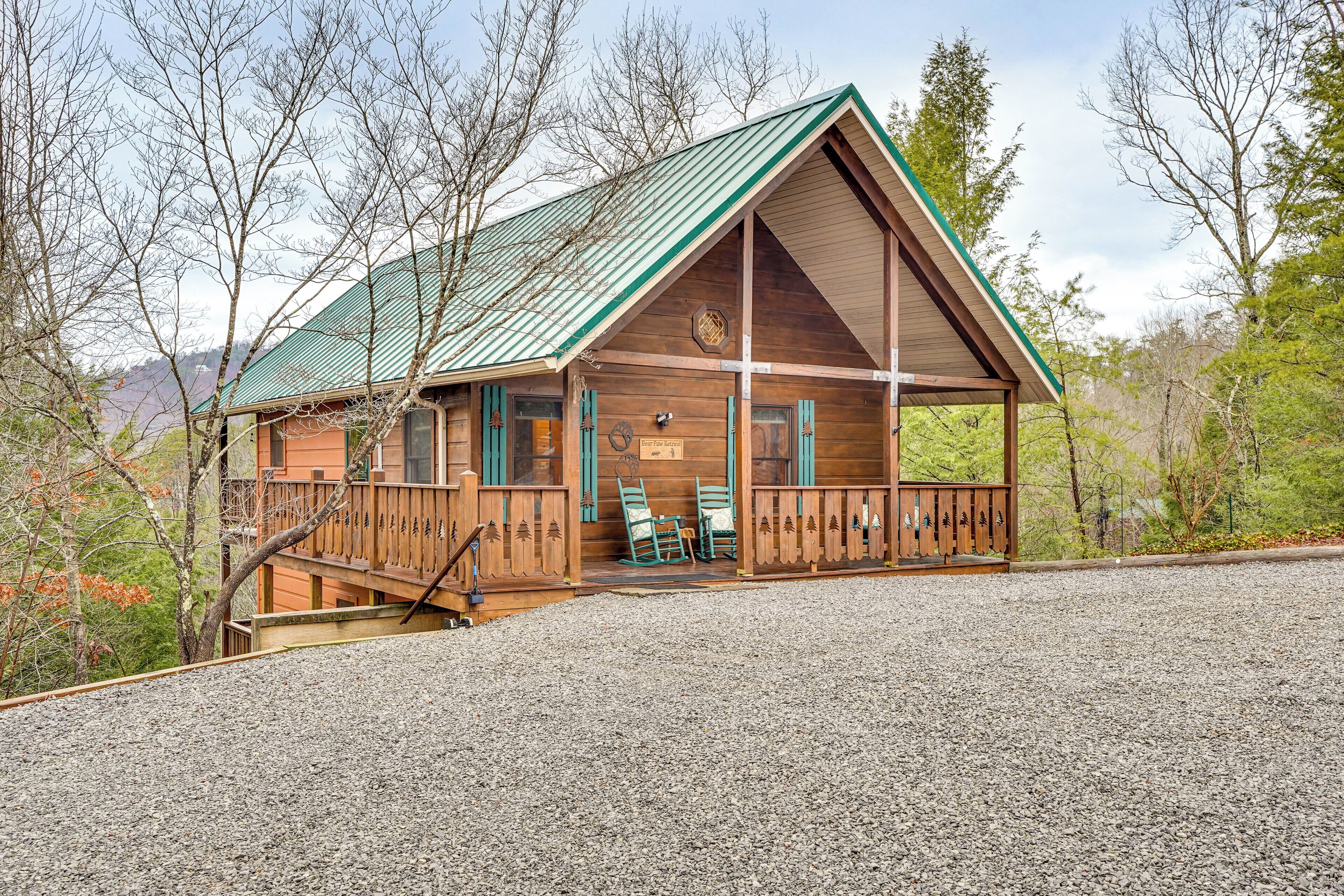 A wooden cabin with a green roof, a porch with chairs, and a gravel driveway.