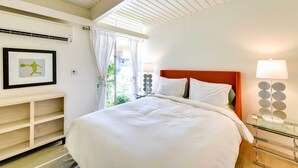 Un lit bien fait, avec du linge de lit blanc, une tête de lit en bois et une table de chevet en verre surmontée d’une lampe au design moderne.
