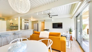 Une cuisine moderne avec des meubles blancs, un coin repas avec une table et des chaises blanches, et un salon avec un canapé jaune et un téléviseur fixé au mur.
