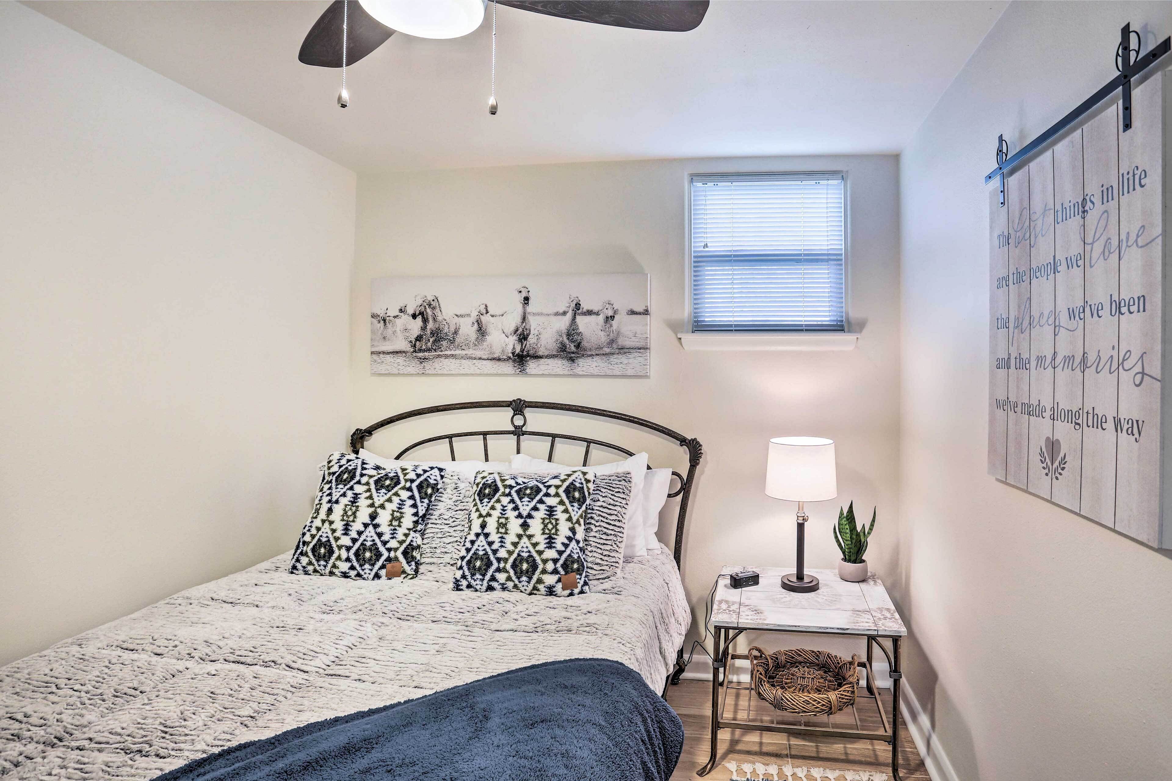 A bedroom with a bed, a ceiling fan, a window, a wall art piece, a small table with a lamp, and a wall-mounted shelf.