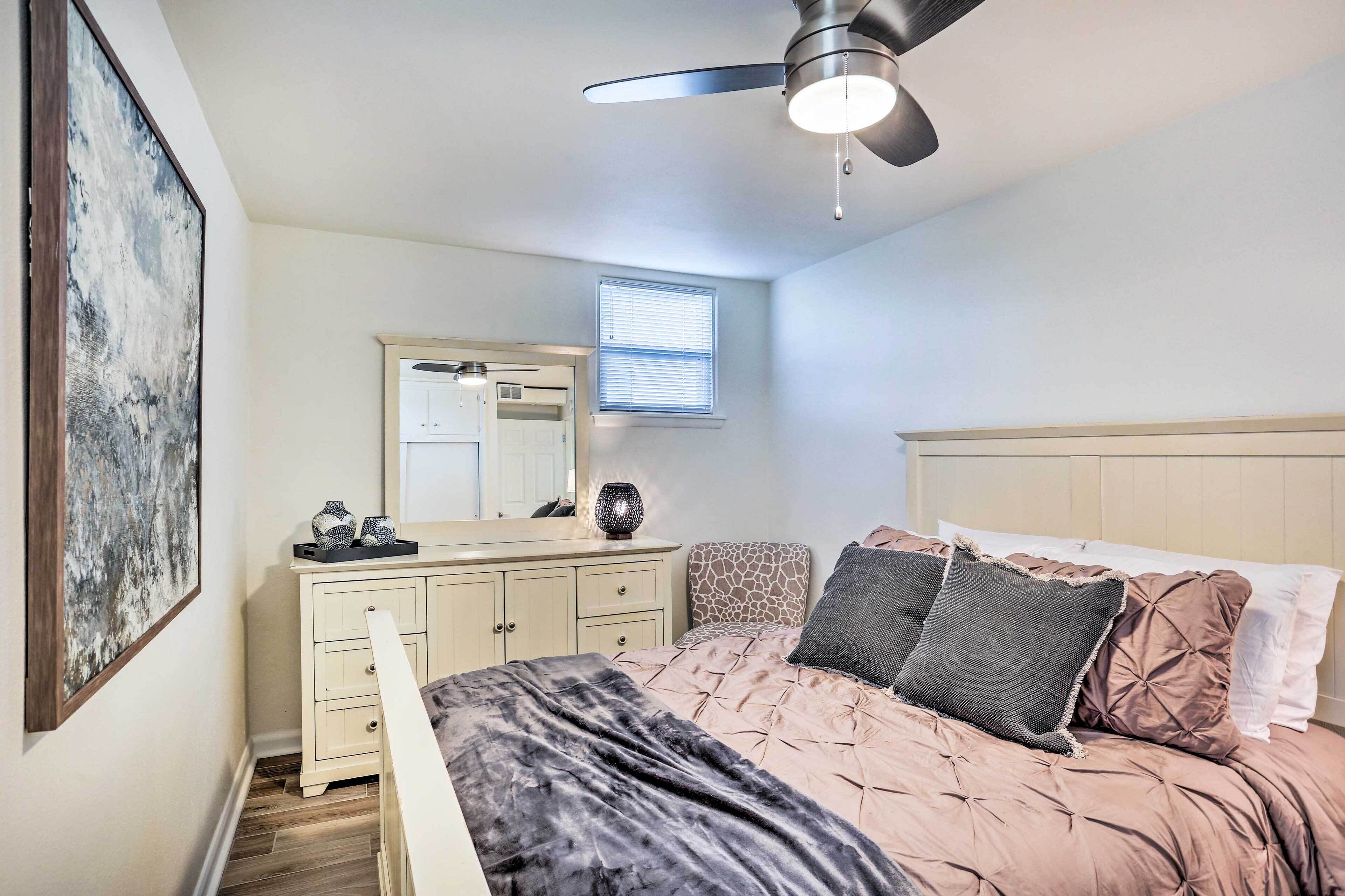 Un dormitorio con cama, cómoda, espejo, ventilador de techo y ventana con persianas.